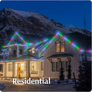 A two-story house with colorful holiday lights on the roof, situated in a snowy area with mountains in the background. The word "Residential" is written at the bottom.