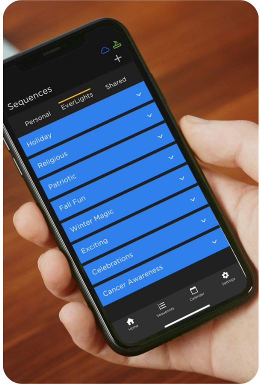 A smartphone screen displays a lighting control app with event category options such as Holiday, Patriotic, Winter Magic, and Cancer Awareness.